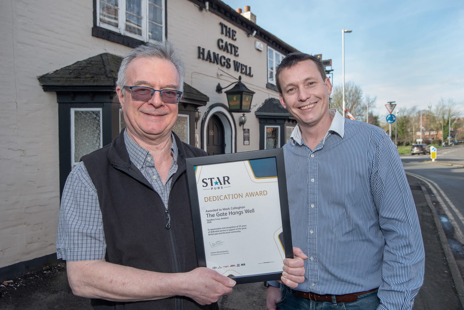 Two men standing outside The Gate Hangs Well Pub holding a certificate